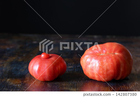 Two ripe red tomatoes, one with unusual natural growth: small tomato has developed on top of larger tomato, creating strikingly irregular appearance, on rustic wooden table against dark background Two ripe red tomatoes, one with unusual natural growth: small tomato has developed on top of larger tomato, creating strikingly irregular appearance, on rustic wooden table against dark background 142807380