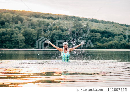 Cold water swimming for elderly women. Senior sporty women splashing in the water, standing in lake during cold evening, arms in the air. Cold water swimming for elderly women. Senior sporty women splashing in the water, standing in lake during cold evening, arms in the air. 142807535