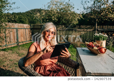 Beautiful older women is sitting garden, holding tablet and scrolling. Beautiful older women is sitting garden, holding tablet and scrolling. 142807539