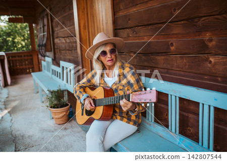 Beautiful older woman is sitting in front of her cabin, playing on guitar. 142807544