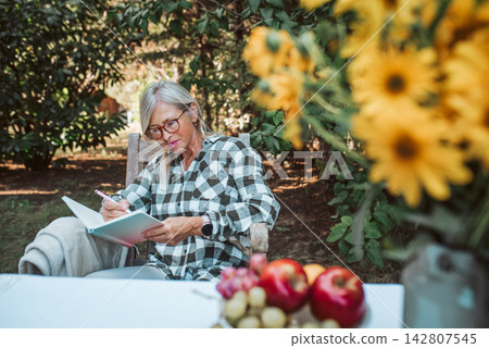 Beautiful older women is sitting in the garden and writing in her diary. Female senior is keeping daily journal, writing down thoughts and feelings. Beautiful older women is sitting in the garden and writing in her diary. Female senior is keeping daily journal, writing down thoughts and feelings. 142807545