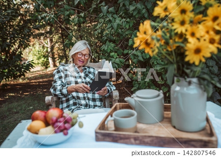 Beautiful older women is sitting in rocking chair in the garden and reading book. Female senior is enjoying a peaceful moment at home, savoring her freedom. Beautiful older women is sitting in rocking chair in the garden and reading book. Female senior is enjoying a peaceful moment at home, savoring her freedom. 142807546