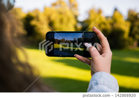 Person photographing a sunlit park with a smartphone, capturing green grass and autumn trees on the screen, creating a modern outdoor lifestyle scene about mobile photography and social media content. 142808920