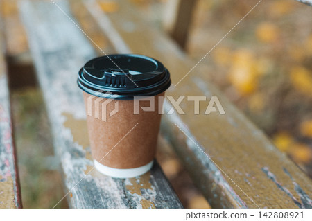 Disposable coffee cup with black lid on a weathered painted wooden bench, creating a cozy outdoor cafe scene with warm seasonal colors. Coffee-to-go. Disposable coffee cup with black lid on a weathered painted wooden bench, creating a cozy outdoor cafe scene with warm seasonal colors. Coffee-to-go. 142808921