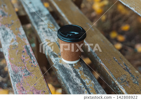 Disposable coffee cup with black lid on a weathered painted wooden bench, creating a cozy outdoor cafe scene with warm seasonal colors. Coffee-to-go. Disposable coffee cup with black lid on a weathered painted wooden bench, creating a cozy outdoor cafe scene with warm seasonal colors. Coffee-to-go. 142808922