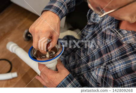 An elderly man focuses on assembling plumbing components for the kitchen and bathroom on a wooden floor. An elderly man focuses on assembling plumbing components for the kitchen and bathroom on a wooden floor. 142809268