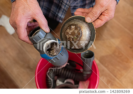 A hand holds a dirty vacuum filter above a red bucket filled with debris and vacuum parts, highlighting the cleanup process in a contemporary kitchen environment. A hand holds a dirty vacuum filter above a red bucket filled with debris and vacuum parts, highlighting the cleanup process in a contemporary kitchen environment. 142809269