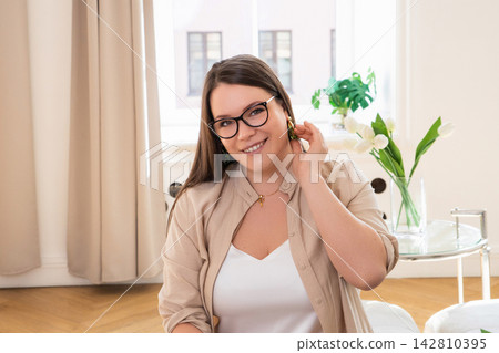 Young woman wearing eyeglasses and earrings posing in bright room. Smiling girl looking at camera indoor. Fashionable lifestyle portrait in modern apartment with white tulip flower. Young woman wearing eyeglasses and earrings posing in bright room. Smiling girl looking at camera indoor. Fashionable lifestyle portrait in modern apartment with white tulip flower. 142810395