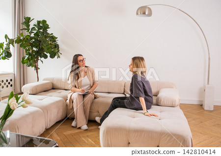 Psychologist talking to woman patient sitting on sofa during session. Therapist taking notes during counseling appointment. Mental health support and professional emotional guidance concept. Psychologist talking to woman patient sitting on sofa during session. Therapist taking notes during counseling appointment. Mental health support and professional emotional guidance concept. 142810414