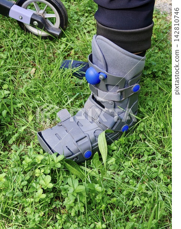 A person wearing an orthopedic boot and using a walker walks across a green lawn. A person wearing an orthopedic boot and using a walker walks across a green lawn. 142810746