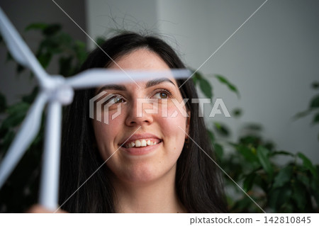 Smiling woman looks past a small wind turbine model in a leafy indoor setting. Clean energy renewable power and female representation 142810845