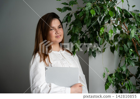 Female freelancer holding a tablet beside a leafy houseplant in a bright home interior. Remote work personal branding and freelance career 142810855