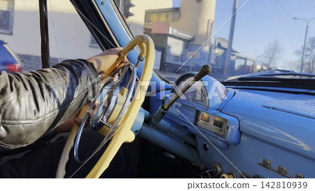 Male hands holds steering wheel while driving retro vehicle at urban street. Man riding on blue vintage auto at city road on sunny day. Driver moving in his old auto on route. Slow motion Male hands holds steering wheel while driving retro vehicle at urban street. Man riding on blue vintage auto at city road on sunny day. Driver moving in his old auto on route. Slow motion 142810939