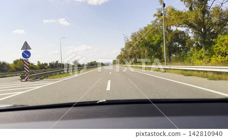 POV from moving car at sunny summer day. Through the windshield white lane marking runs steadily along asphalt road, creating a clear sense of speed, rhythm and open daytime travel POV from moving car at sunny summer day. Through the windshield white lane marking runs steadily along asphalt road, creating a clear sense of speed, rhythm and open daytime travel 142810940