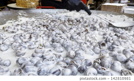 Male hand in glove stirs snails shells in creamy sauce with long spoon during cooking process in restaurant kitchen. The snails simmer gently on metal cooking surface coated in rich sauce. Slow motion Male hand in glove stirs snails shells in creamy sauce with long spoon during cooking process in restaurant kitchen. The snails simmer gently on metal cooking surface coated in rich sauce. Slow motion 142810981