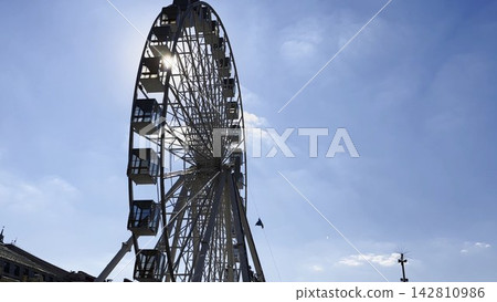 View of a ferris wheel against a clear blue sky with sunlight at background. The sun rays pass through the structure, creating bright highlights and a vibrant summer atmosphere around the attraction View of a ferris wheel against a clear blue sky with sunlight at background. The sun rays pass through the structure, creating bright highlights and a vibrant summer atmosphere around the attraction 142810986