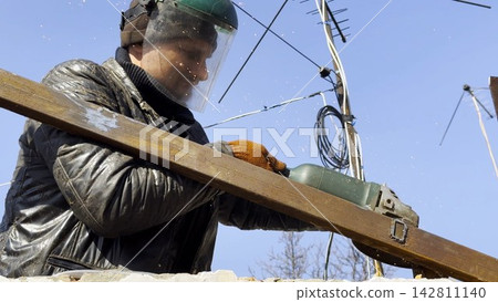 Young mechanic in protective gloves polishing iron using electric grinder wheel at construction. Professional repairman grinding metal details with circular saw on house at sunny day. Slow mo 142811140