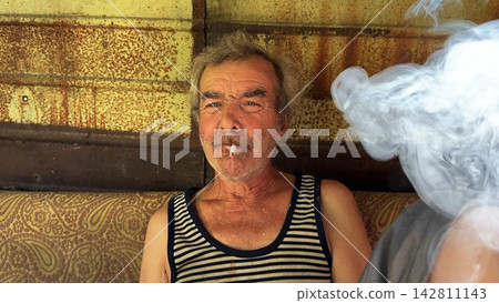 Portrait of homeless adult man sitting on worn couch placed and smoking outdoor. The setting underscores neglect and impermanence, presenting a raw and unfiltered glimpse of difficult living situation Portrait of homeless adult man sitting on worn couch placed and smoking outdoor. The setting underscores neglect and impermanence, presenting a raw and unfiltered glimpse of difficult living situation 142811143