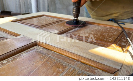 Male hands use electric polishing machine to smooth and refine wooden door surface. The rotating pad carefully treats wood, enhancing texture and finish during detailed restoration or renovation work Male hands use electric polishing machine to smooth and refine wooden door surface. The rotating pad carefully treats wood, enhancing texture and finish during detailed restoration or renovation work 142811148