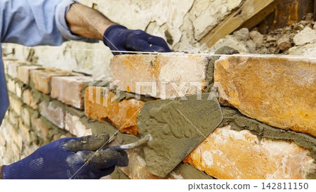 Adult male hands in protective gloves apply cement with a trowel and place bricks to build brick wall for house. The worker carefully spreads mortar and sets each brick during the construction process Adult male hands in protective gloves apply cement with a trowel and place bricks to build brick wall for house. The worker carefully spreads mortar and sets each brick during the construction process 142811150