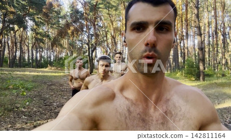 Group of young muscular athletes running at forest path. Active strong men jogging at sunny woodland. Male shirtless sportsmen training at summer nature. Concept of active and healthy lifestyle. POV 142811164