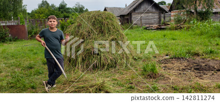Young boy posing with a hay fork next to a large pile of freshly cut grass in a rural village setting, symbolizing childhood, farm life, agriculture, and summer work outdoors. Young boy posing with a hay fork next to a large pile of freshly cut grass in a rural village setting, symbolizing childhood, farm life, agriculture, and summer work outdoors. 142811274