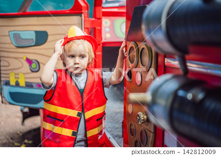 Little boy dressed as firefighter playing in a red toy fire truck at an outdoor playground with helmet safety vest pretend rescue game childhood dreams role play and side view. Little boy dressed as firefighter playing in a red toy fire truck at an outdoor playground with helmet safety vest pretend rescue game childhood dreams role play and side view. 142812009