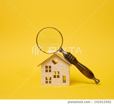 Magnifying glass with a wooden handle lying on a decorative toy house on a bright yellow background. Magnifying glass with a wooden handle lying on a decorative toy house on a bright yellow background. 142812082