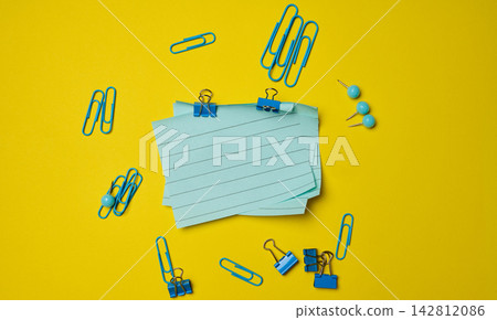 Blue stationery supplies a stack of sheets of paper, paper clips, and pushpins on a yellow background Blue stationery supplies a stack of sheets of paper, paper clips, and pushpins on a yellow background 142812086