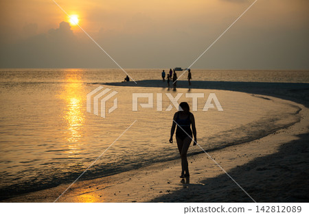 Silhouette of a woman in a swimsuit walking on a sandy beach at sunset, Omadhoo, Maldives. Golden hour reflection on the ocean water. Summer vacation travel and sea concept Silhouette of a woman in a swimsuit walking on a sandy beach at sunset, Omadhoo, Maldives. Golden hour reflection on the ocean water. Summer vacation travel and sea concept 142812089