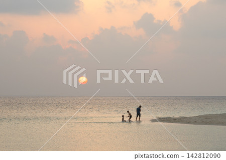 Silhouette of a man and children playing in the shallow ocean water at sunset, Maldives. Family vacation at the sea beach with orange sun on the horizon. Copy space for text Silhouette of a man and children playing in the shallow ocean water at sunset, Maldives. Family vacation at the sea beach with orange sun on the horizon. Copy space for text 142812090