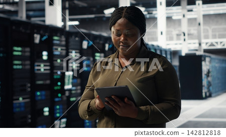 Technician using tablet in data center, integrating new technologies and systems. Employee using device to oversee server hub, ensuring compliance with industry standards, camera B Technician using tablet in data center, integrating new technologies and systems. Employee using device to oversee server hub, ensuring compliance with industry standards, camera B 142812818