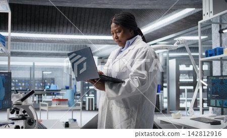 African American woman scientist works in a modern laboratory, conducting experiments with advanced equipment and a laptop. Molecular biology, biotechnology and healthcare innovation. African American woman scientist works in a modern laboratory, conducting experiments with advanced equipment and a laptop. Molecular biology, biotechnology and healthcare innovation. 142812916