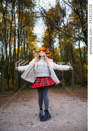 Happy young woman wearing a maple leaf wreath enjoys an autumn park with open arms. Fall lifestyle, colorful foliage, outdoor leisure, wellness and seasonal nature during a weekend walk. Happy young woman wearing a maple leaf wreath enjoys an autumn park with open arms. Fall lifestyle, colorful foliage, outdoor leisure, wellness and seasonal nature during a weekend walk. 142813528