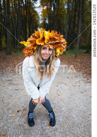 Happy young woman wearing a maple leaf wreath enjoys an autumn park with open arms. Fall lifestyle, colorful foliage, outdoor leisure, wellness and seasonal nature during a weekend walk. Happy young woman wearing a maple leaf wreath enjoys an autumn park with open arms. Fall lifestyle, colorful foliage, outdoor leisure, wellness and seasonal nature during a weekend walk. 142813529