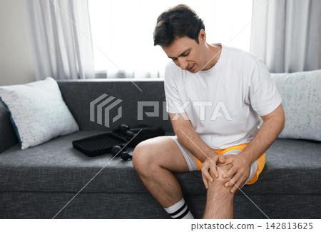 Man with knee pain and muscle tension prepares for recovery and rehabilitation. He sits near a percussion massage device for physical therapy and self care relief. Man with knee pain and muscle tension prepares for recovery and rehabilitation. He sits near a percussion massage device for physical therapy and self care relief. 142813625