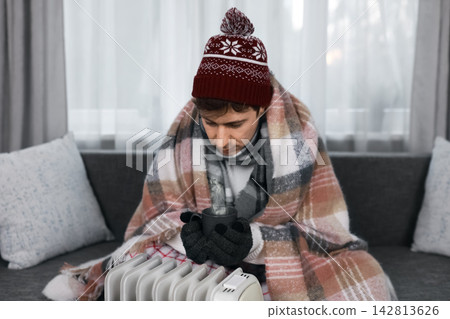 Freezing man indoors during winter frost battles a heating problem. He relies on a heater and hot drink to beat cold, energy crisis, high costs, and low temperature. Freezing man indoors during winter frost battles a heating problem. He relies on a heater and hot drink to beat cold, energy crisis, high costs, and low temperature. 142813626
