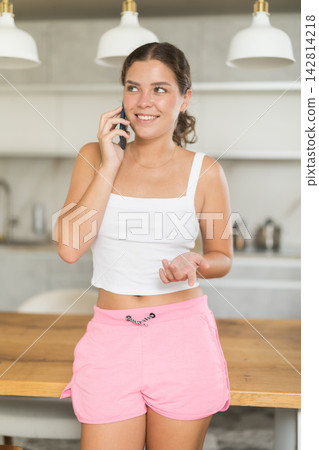 Expressive young girl wearing shorts talking on smartphone while standing in kitchen Expressive young girl wearing shorts talking on smartphone while standing in kitchen 142814218