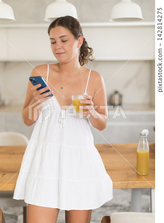Girl with smartphone drinks juice in kitchen Girl with smartphone drinks juice in kitchen 142814275
