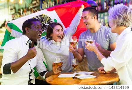 Friends gathered together to watch soccer football championship and celebrating with glasses of beer. Girl holding flag of Mexico 142814399