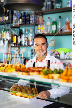 Bartender standing at counter in bar, selling fresh sandwiches pinchos to client Bartender standing at counter in bar, selling fresh sandwiches pinchos to client 142814482