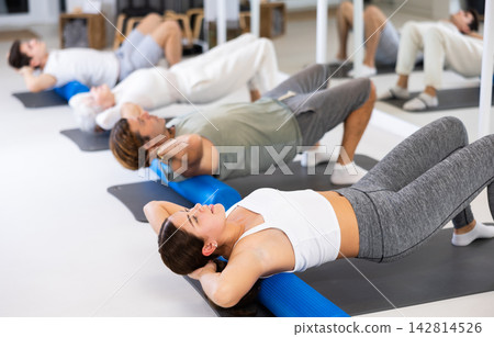 Young positive female performing set of exercises with pilates roller during group class in fitness studio. Young positive female performing set of exercises with pilates roller during group class in fitness studio. 142814526