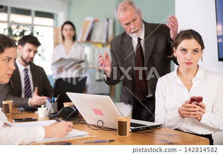 Strict mature man boss scolding girl office worker for scrolling social media on smartphone during work hours in office 142814582