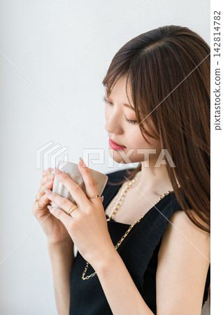 A woman with a serene expression gazes at a cup. An elegant moment of relaxation, adorned with jewelry. A woman with a serene expression gazes at a cup. An elegant moment of relaxation, adorned with jewelry. 142814782