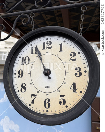 Round wall clock shows time at 10:10 with a black frame, hanging on a decorative structure outdoors against a blue background Round wall clock shows time at 10:10 with a black frame, hanging on a decorative structure outdoors against a blue background 142817798