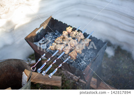 Grilled meat skewers are placed on a charcoal barbecue grill with smoke rising, set in an outdoor environment with natural elements visible Grilled meat skewers are placed on a charcoal barbecue grill with smoke rising, set in an outdoor environment with natural elements visible 142817894