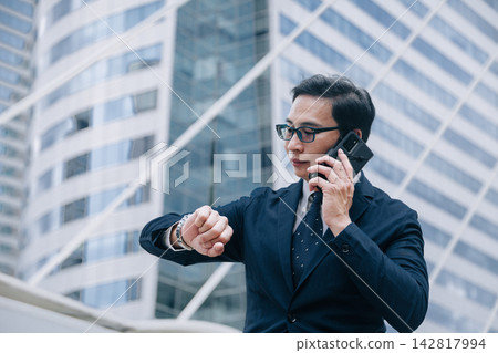 Asian Business man make a Phone Call while look at wrist watch for Appointment Times, Contact meeting Time, Business Hours. Asian Business man make a Phone Call while look at wrist watch for Appointment Times, Contact meeting Time, Business Hours. 142817994