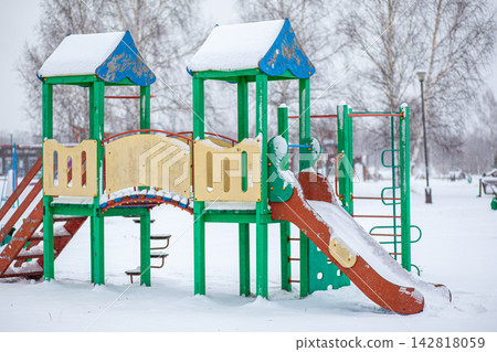 Colorful playground equipment with a slide and climbing structure is covered in snow, set in a winter park with trees in the background Colorful playground equipment with a slide and climbing structure is covered in snow, set in a winter park with trees in the background 142818059