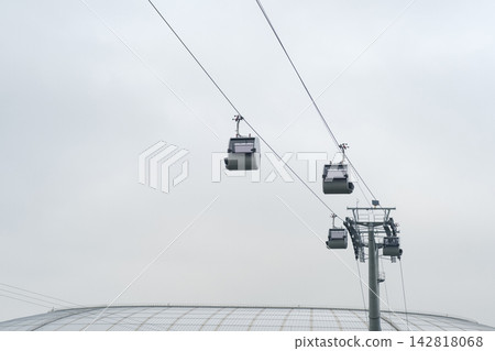 Cable cars travel along overhead lines in an urban area, with a large building structure visible beneath them against a cloudy sky Cable cars travel along overhead lines in an urban area, with a large building structure visible beneath them against a cloudy sky 142818068