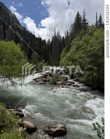 Flowing river with clear water meanders through a landscape of green trees and rocky banks under a partly cloudy sky in the mountains Flowing river with clear water meanders through a landscape of green trees and rocky banks under a partly cloudy sky in the mountains 142818103